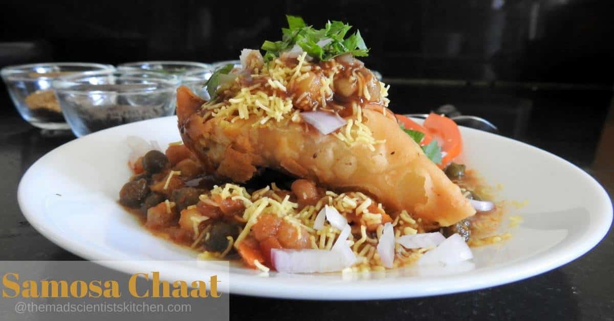 Samosa Chaat is mashed samosa topped with peas or chole masala and topped with tangy chutneys, sev, onion and tomatoes if you wish.