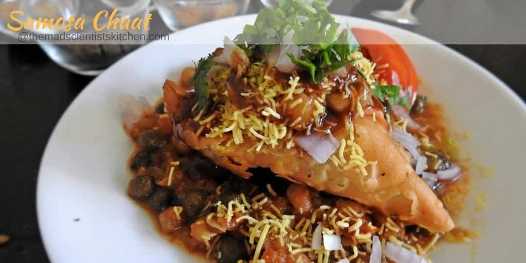 Samosa Chaat is mashed samosa topped with peas or chole masala and topped with tangy chutneys, sev, onion and tomatoes if you wish.